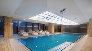 A modern indoor swimming pool with a clear ceiling, lounge chairs, and a view through large windows. - Hilton Garden Inn Istanbul Airport (Istanbul)