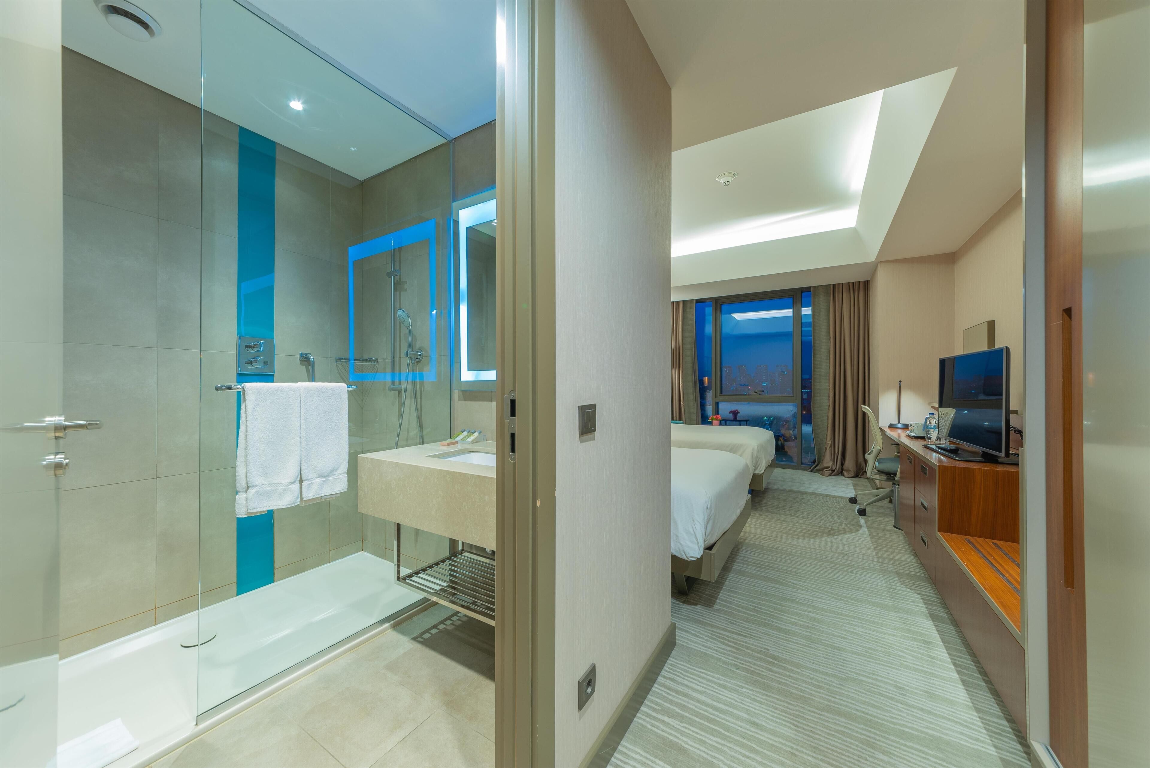 A modern hotel room with a glass-enclosed shower, a separate bathroom, a bed, a desk with a computer, and a television.