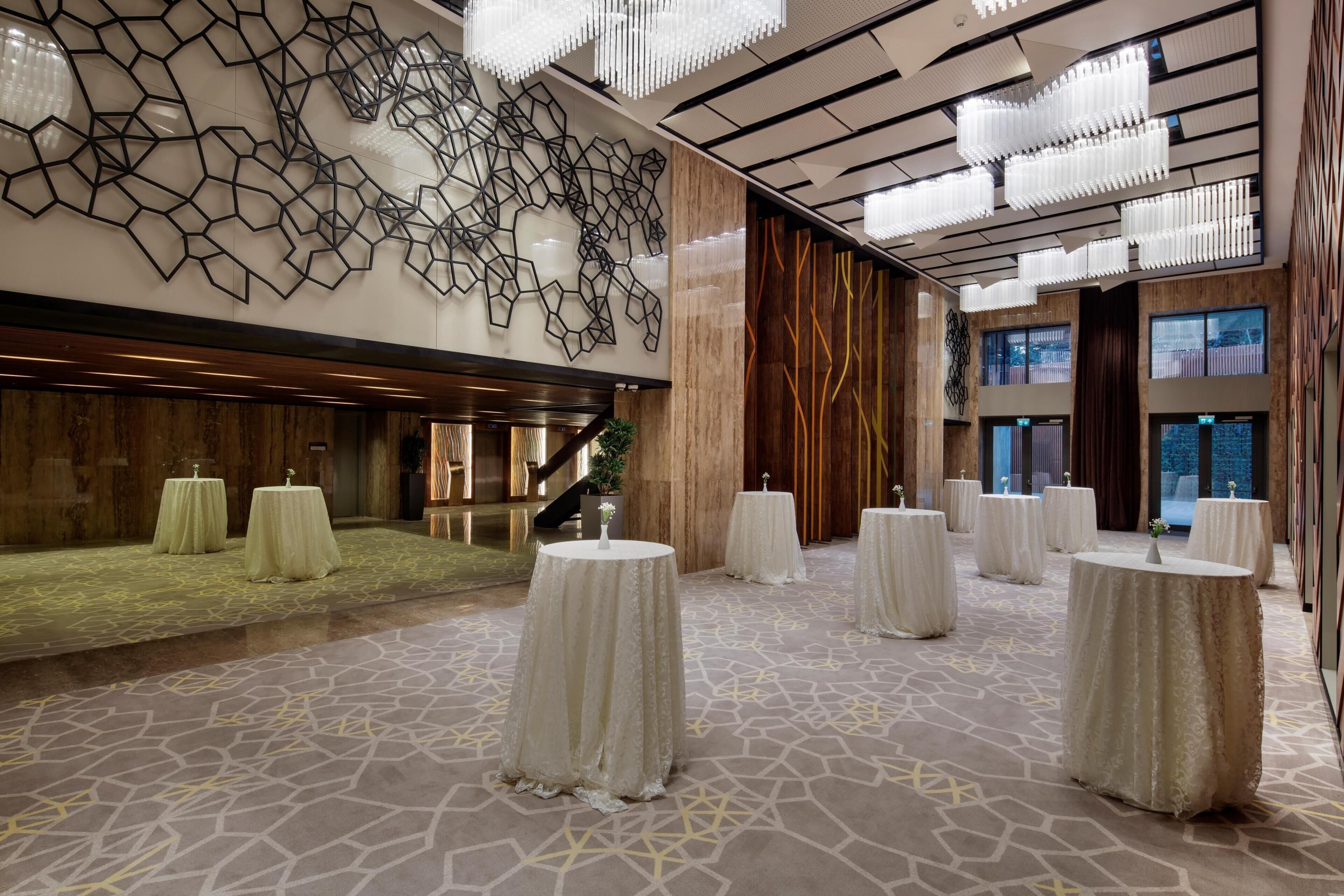 A spacious lobby with a geometric wall design, multiple round tables, and a staircase leading to upper levels.