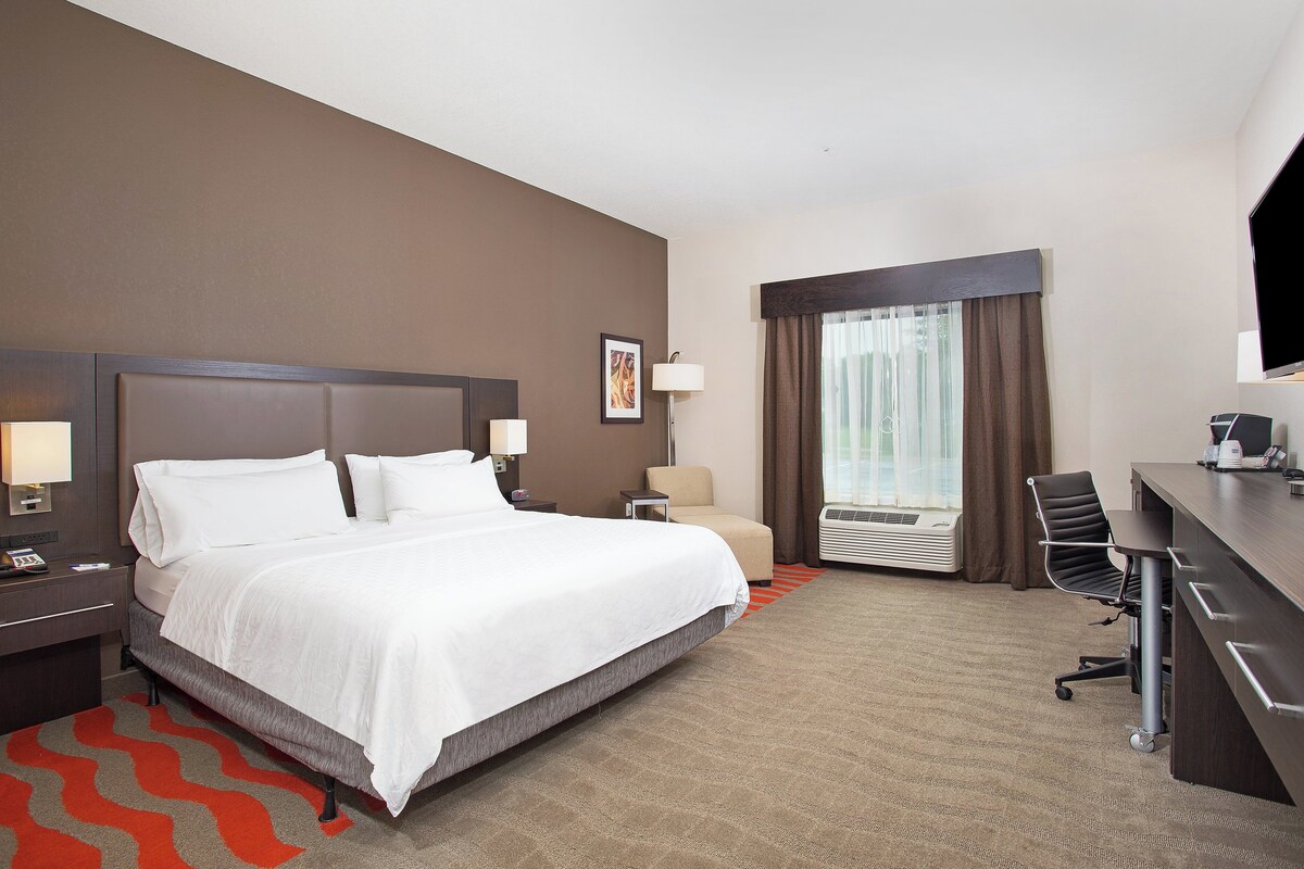 A hotel room with a large bed, a desk, a chair, a lamp, a picture on the wall, and a window with curtains.