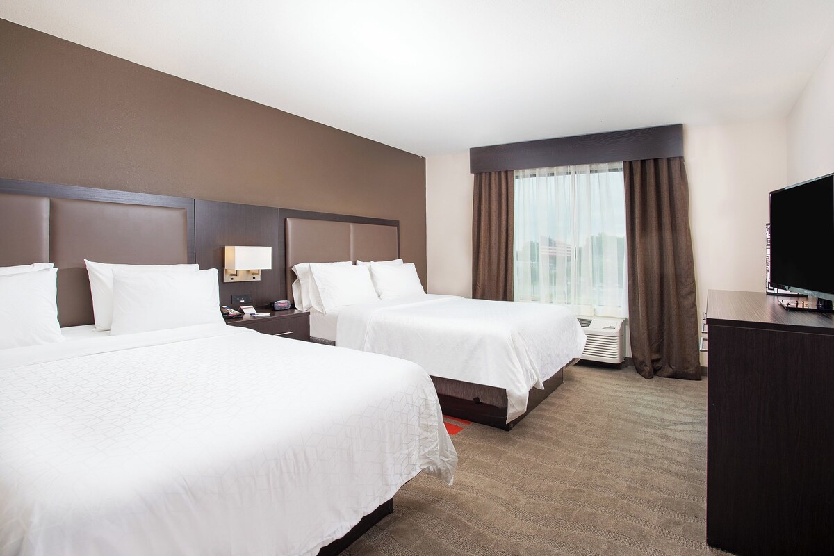 A hotel room with two beds, a flat-screen TV, a desk, and a large window with curtains.