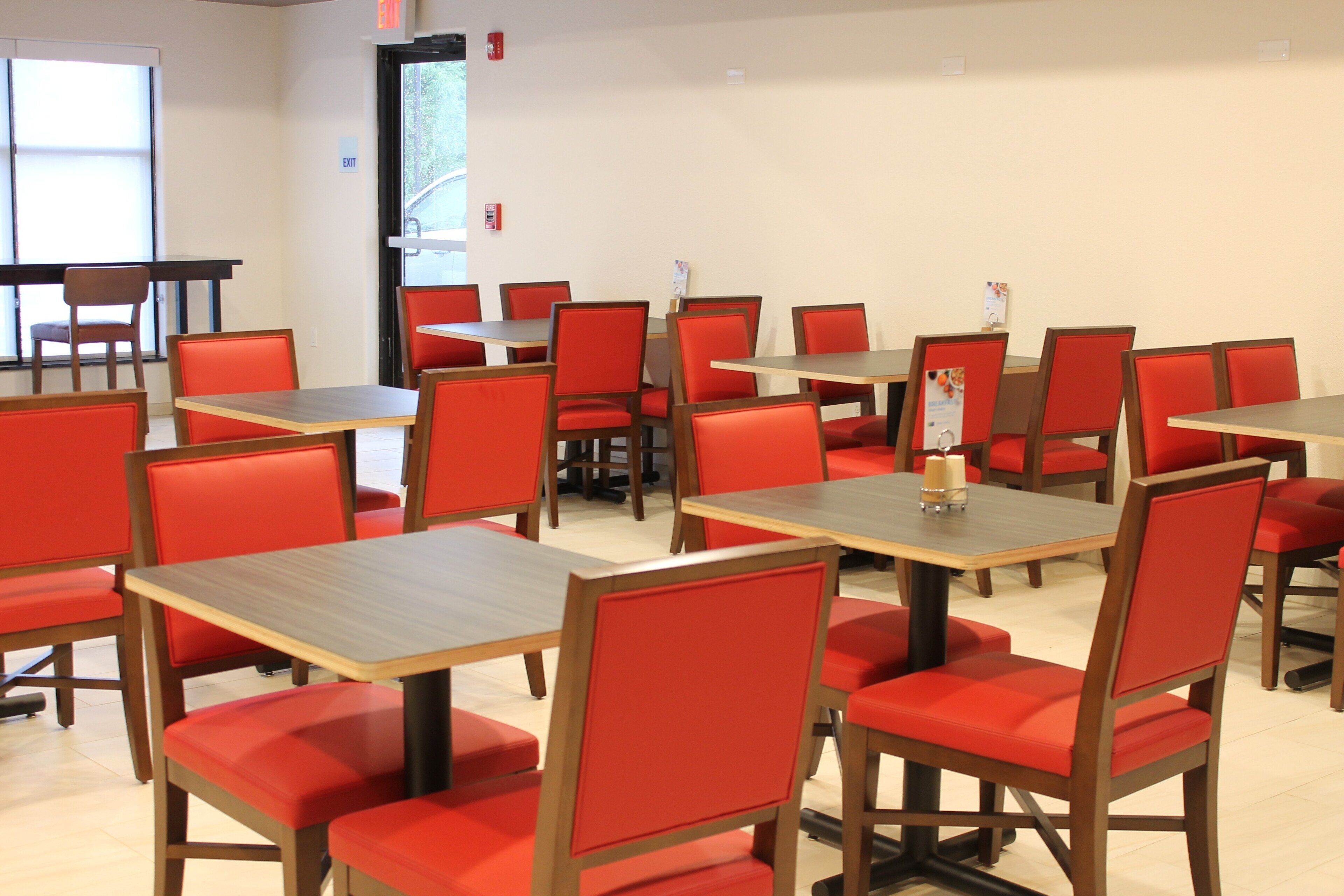 A dining area with multiple tables and red chairs, a fire extinguisher, and an exit sign on the wall.