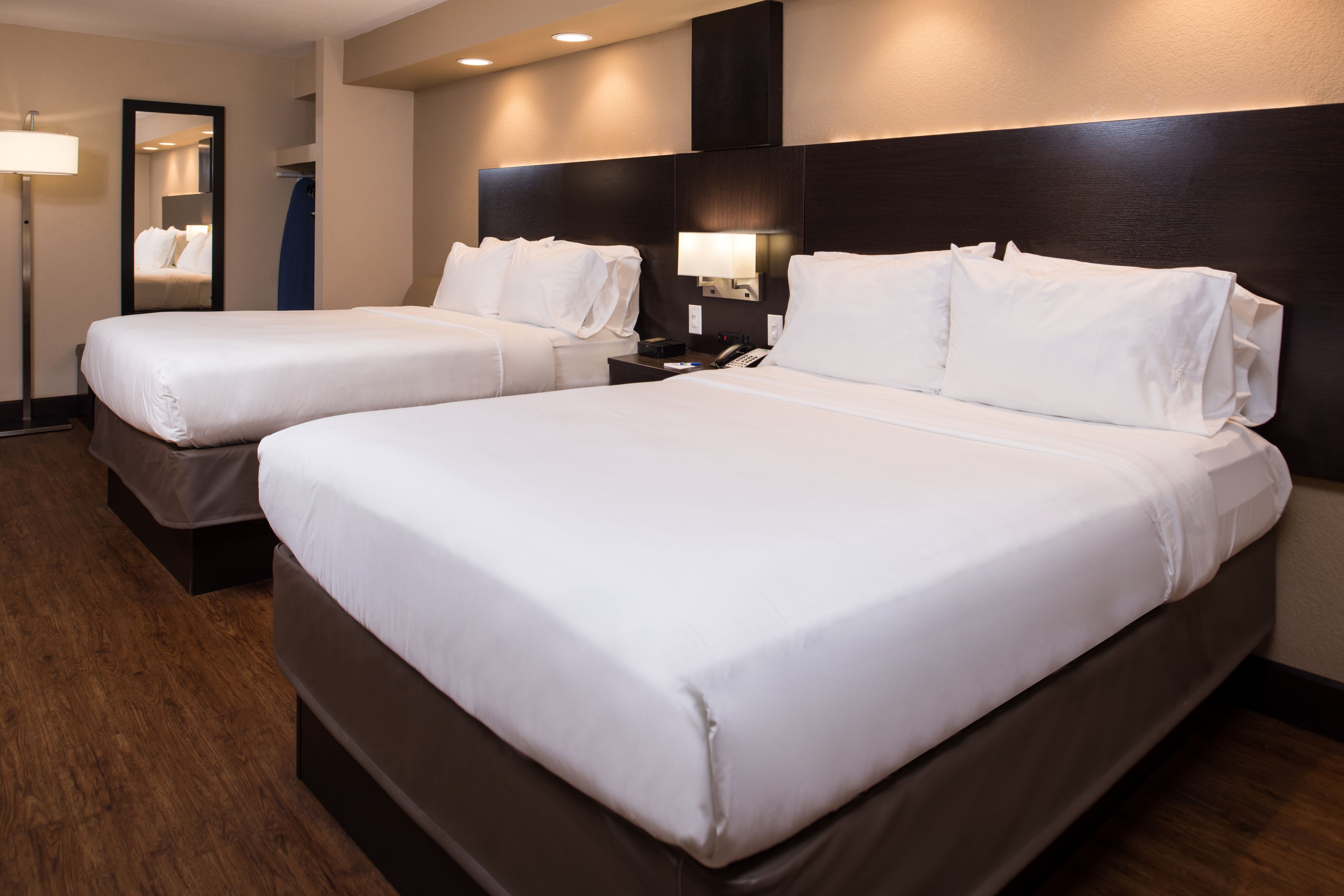 Habitación de hotel con dos camas individuales, una mesita de noche con lámpara, un espejo y un cabecero oscuro.