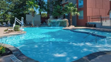 A swimming pool with a waterfall feature, surrounded by palm trees and lounge chairs.