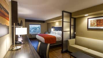 Habitación de hotel con una cama, un escritorio con lámpara, un sofá y una ventana con vistas a árboles.