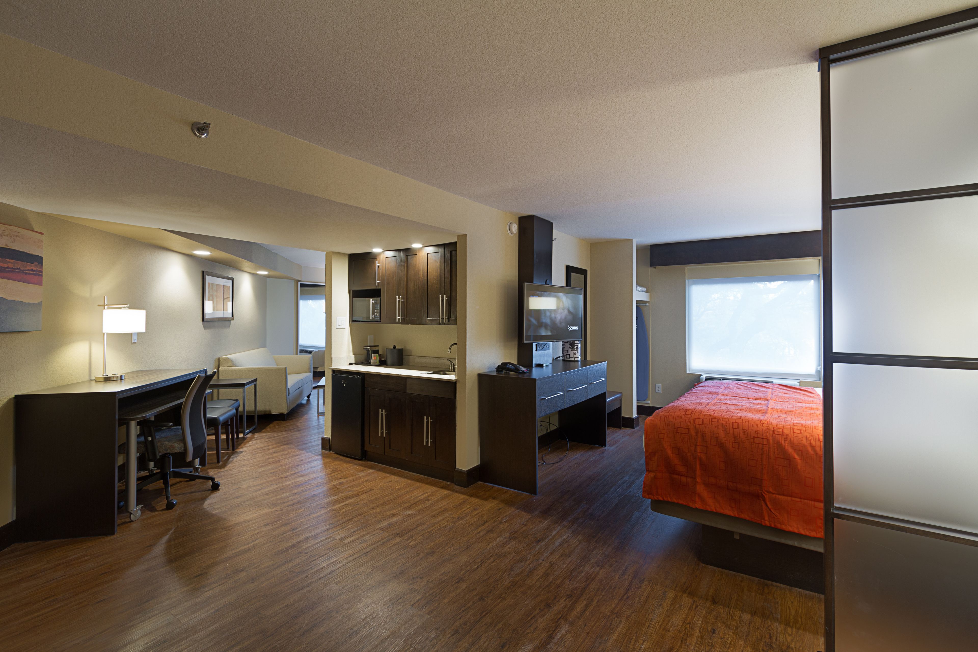 A modern hotel room with a wooden floor, a desk with a chair, a bed with an orange bedspread, a kitchenette with dark cabinets, and a large window providing natural light.