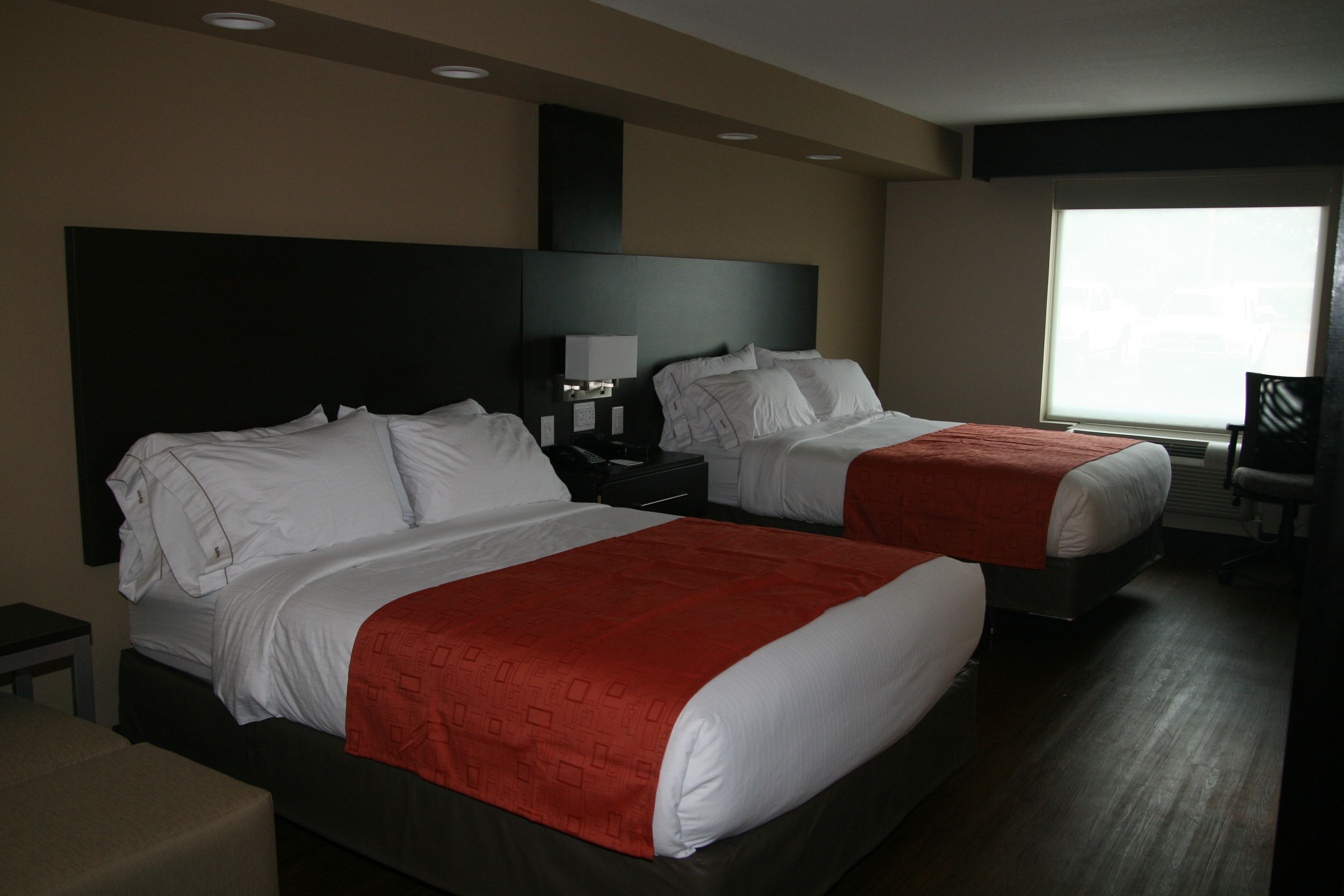 A hotel room with two beds, each with white bedding and red headboards. There is a nightstand with a lamp between the beds, and a window providing natural light.