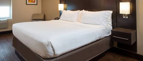Una cama grande con ropa blanca, un cabecero con iluminación integrada, una mesita de noche con lámpara y una silla en una habitación de hotel.