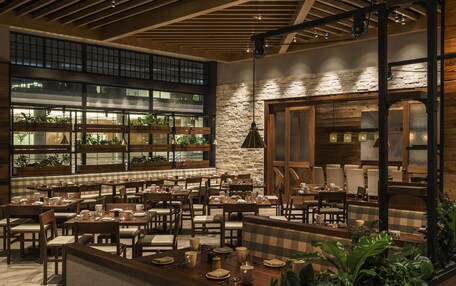 A spacious restaurant with multiple dining tables and booths, large windows with plants, and a stone wall.
