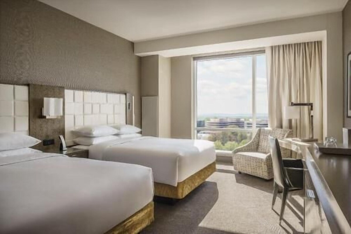 A hotel room with two beds, a desk, a chair, and a large window offering a city view.