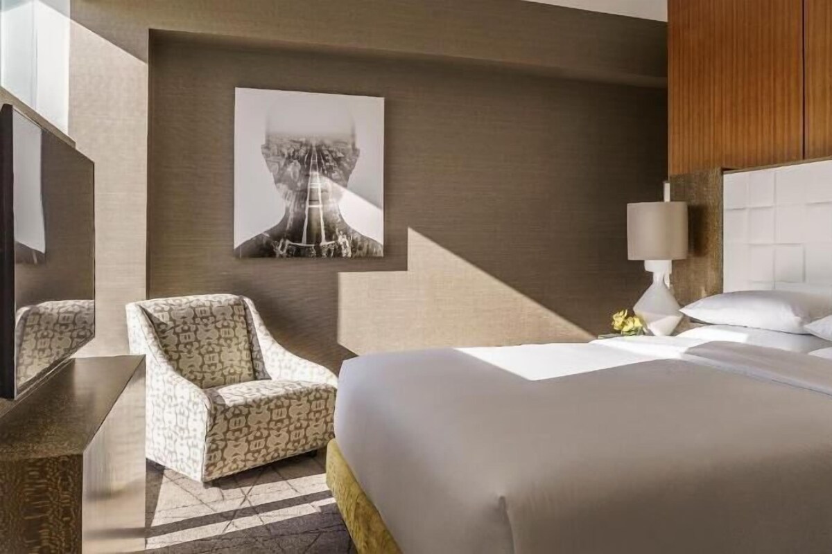 A hotel room with a bed, a chair, a lamp, and a wall-mounted artwork.