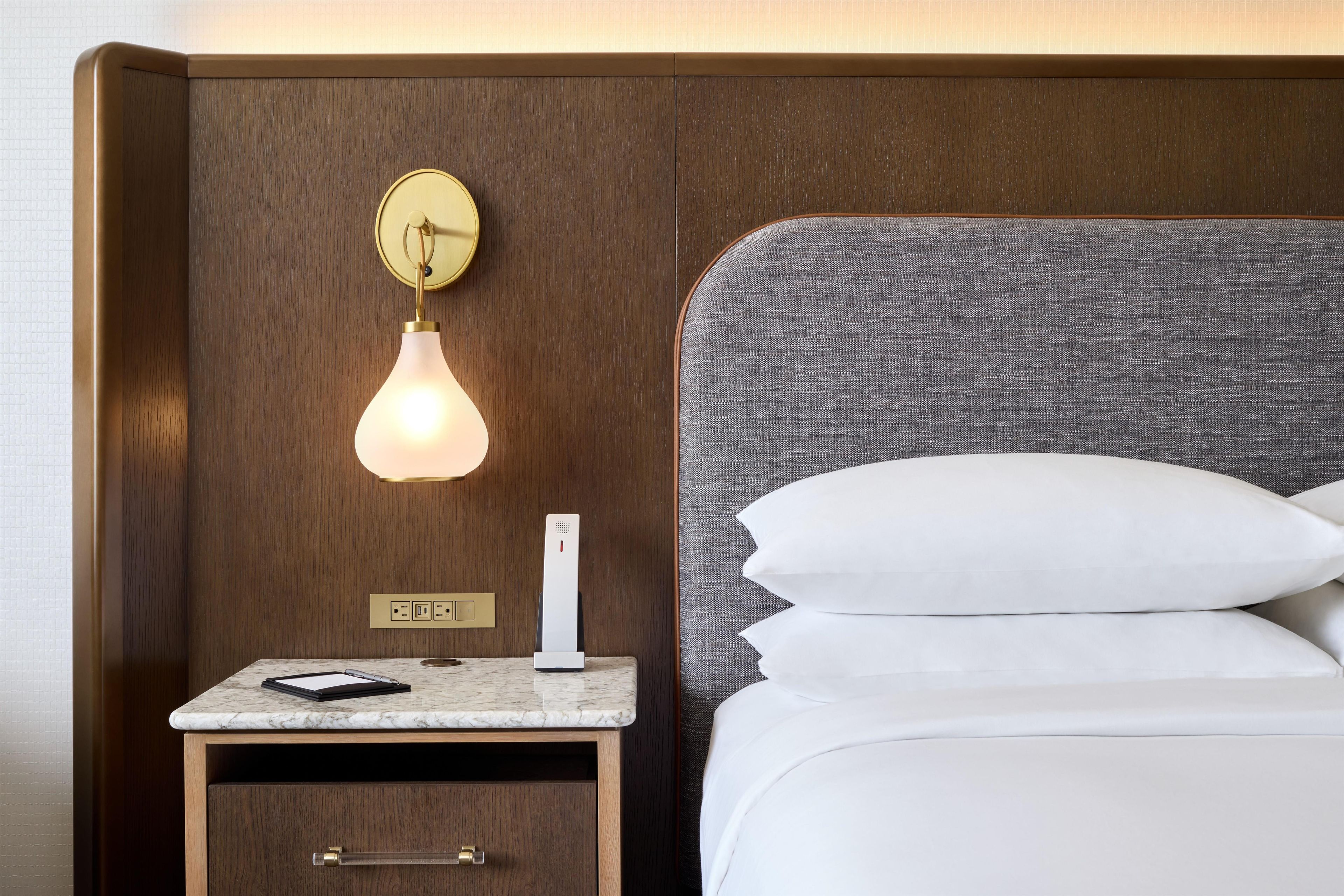 A hotel room with a wooden headboard, a bedside table with a lamp, a phone, and a notepad.