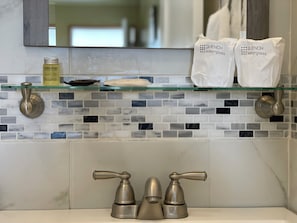 A bathroom with a glass shelf holding toiletries, a bottle of shampoo, and two white bags labeled 'QUENCH waterglass'. - Walla Walla Garden Motel (Walla Walla)