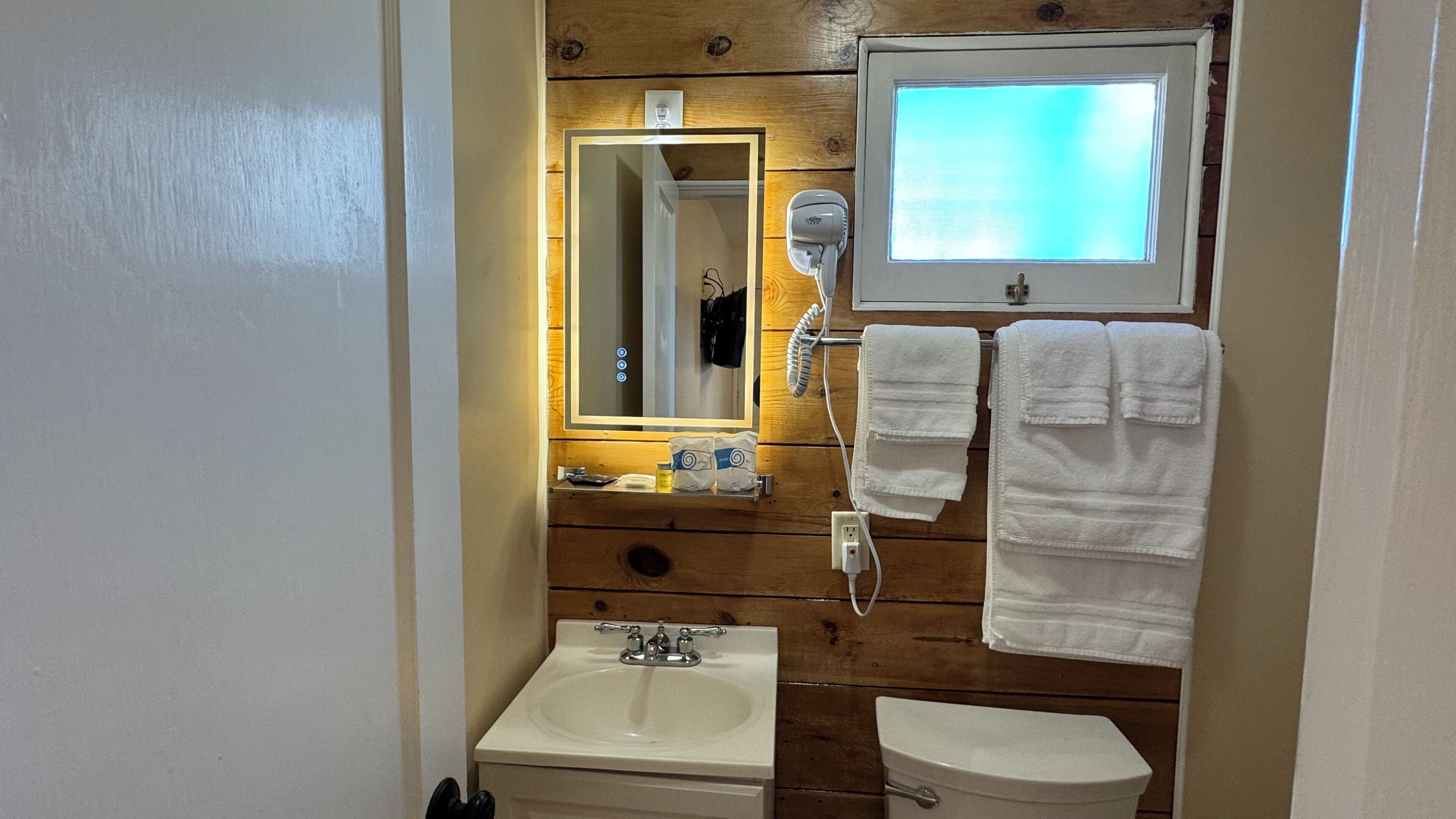 Un baño con pared de madera, un espejo, un secador de pelo, un lavamanos y un inodoro.