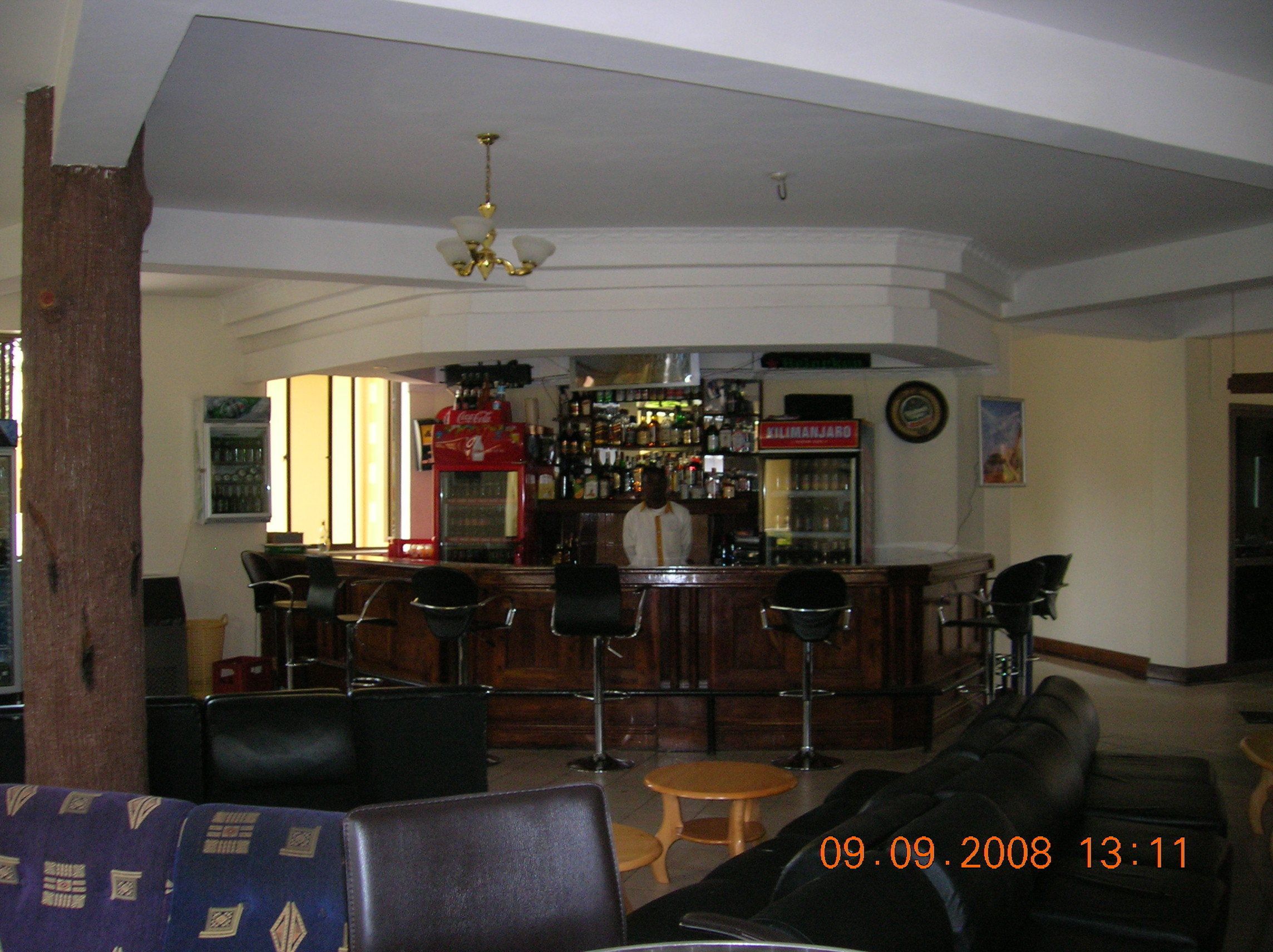 A bar area with a counter, stools, and a refrigerator. There are chairs and a sofa in the foreground.