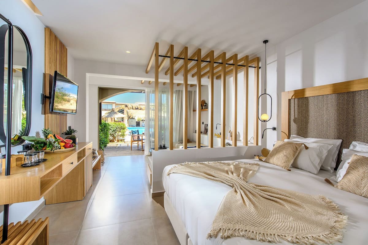 Een moderne hotelkamer met een groot bed, een bureau met spiegel en uitzicht op het zwembad en de tuin.