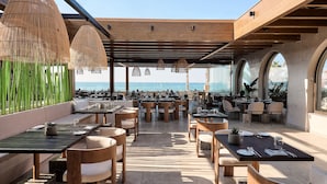 An outdoor dining area with tables and chairs, a view of the sea, and a covered section with hanging lights. - Stella Palace Aqua Park Resort (Hersonissos)