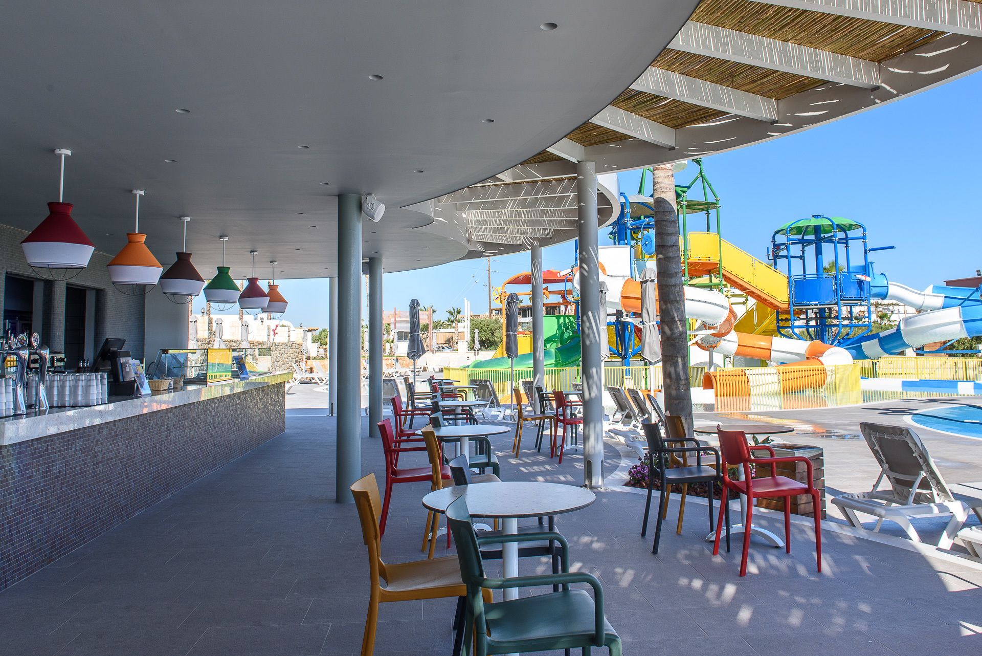 An outdoor seating area with tables and chairs, a bar, and a water park in the background.