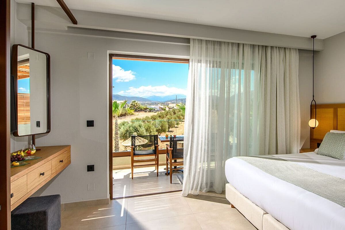Een moderne hotelkamer met een groot bed, een houten hoofdeinde, een bureau met spiegel en een balkon met uitzicht op het landschap.