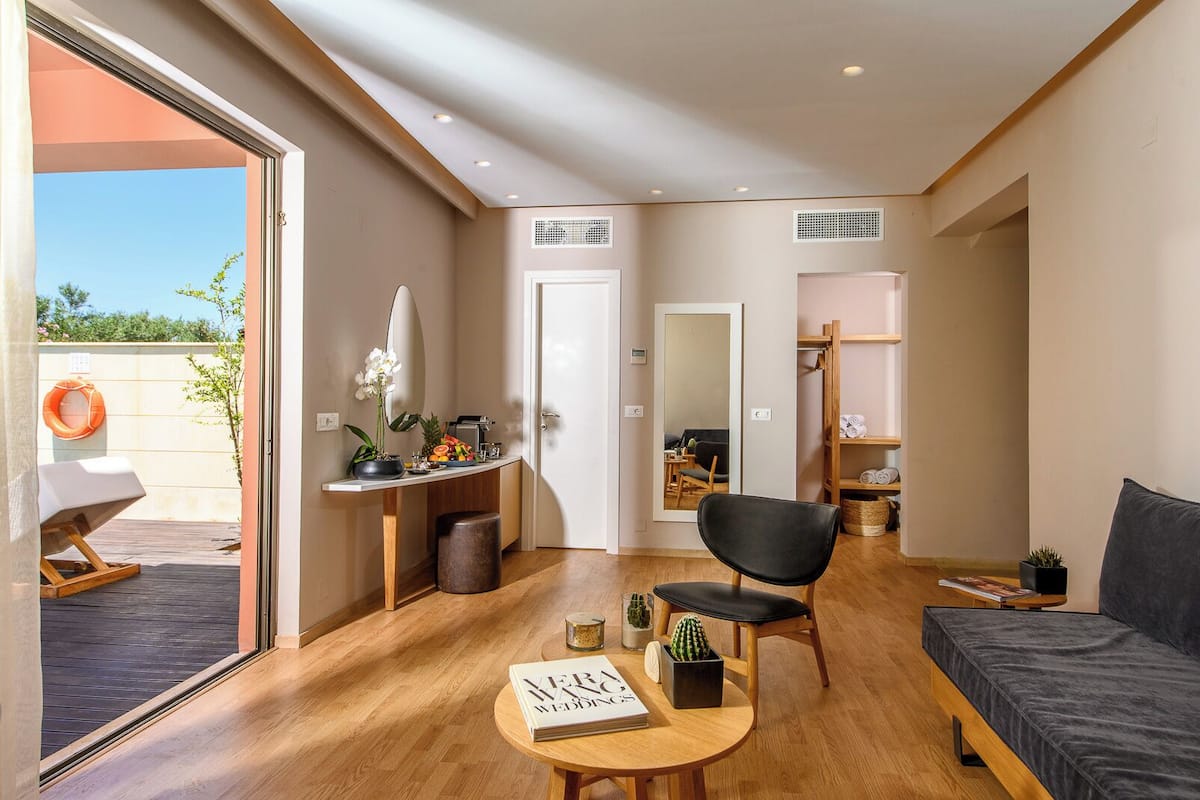 Een moderne woonkamer met een bank, een stoel, een klein tafeltje met een boek en een salontafel. Er is een deur naar een andere kamer, een klein plankje met spullen en een raam met uitzicht op de omgeving.