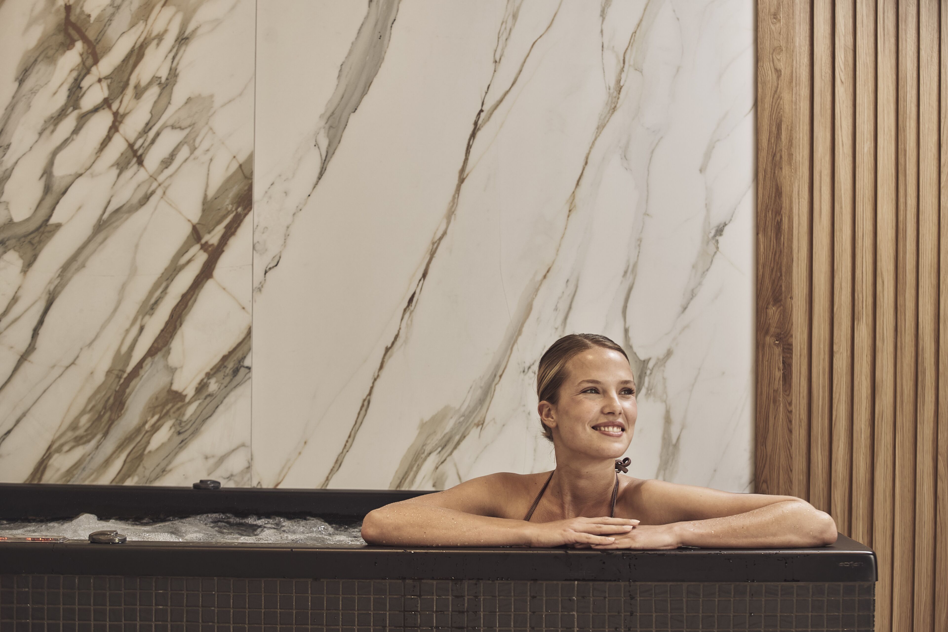 Una persona relajándose en una bañera con una pared de mármol y paneles de madera al fondo.