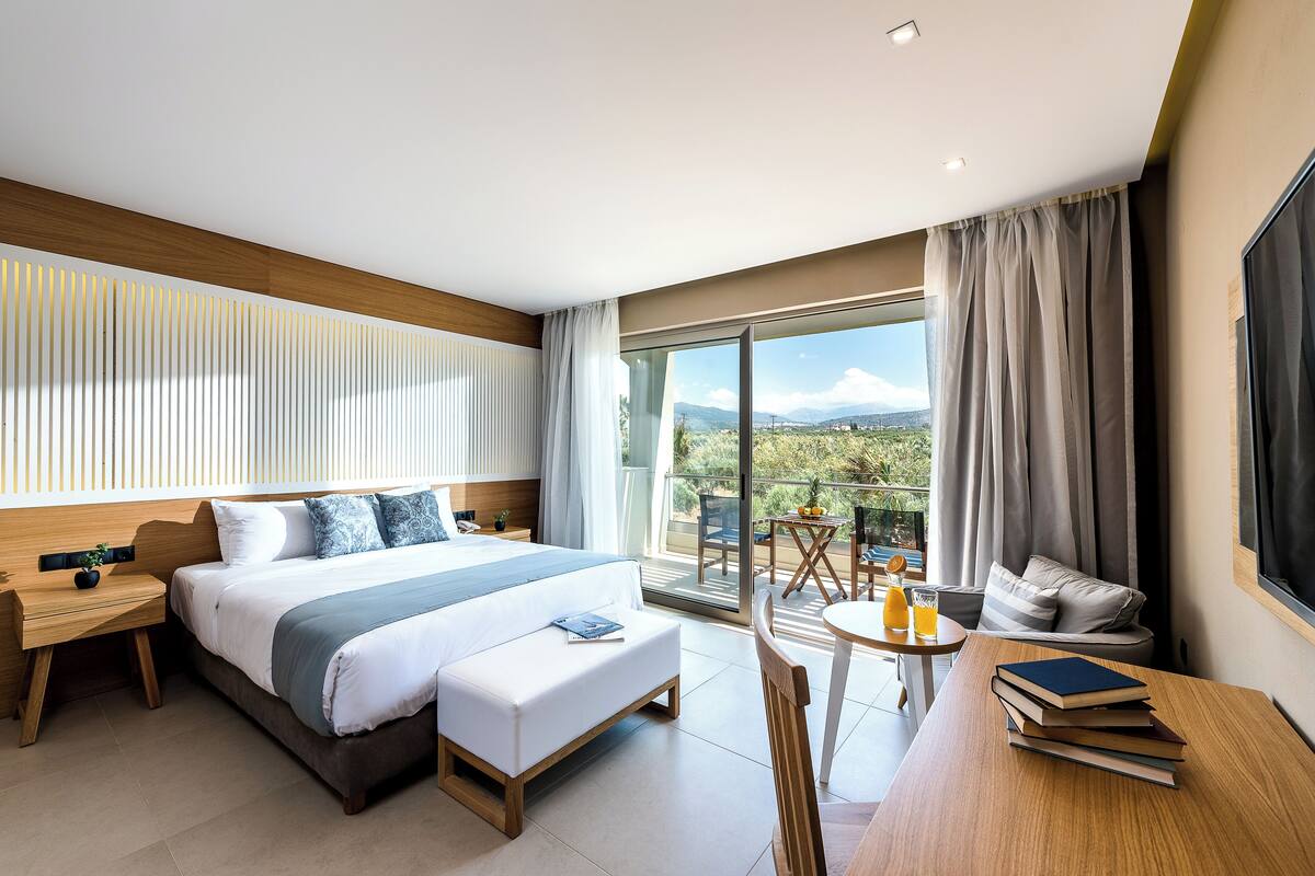 Een moderne hotelkamer met een groot bed, een balkon met tafel en stoelen, en uitzicht op de omgeving.