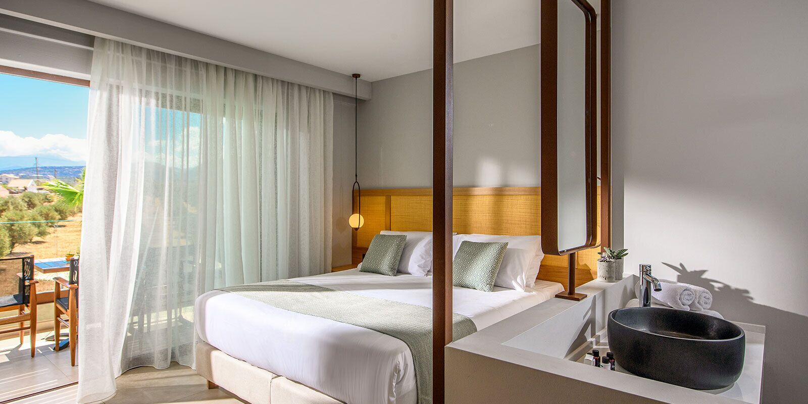 A hotel room with a bed, a bathroom with a sink and a large basin, and a balcony with a view of the countryside.