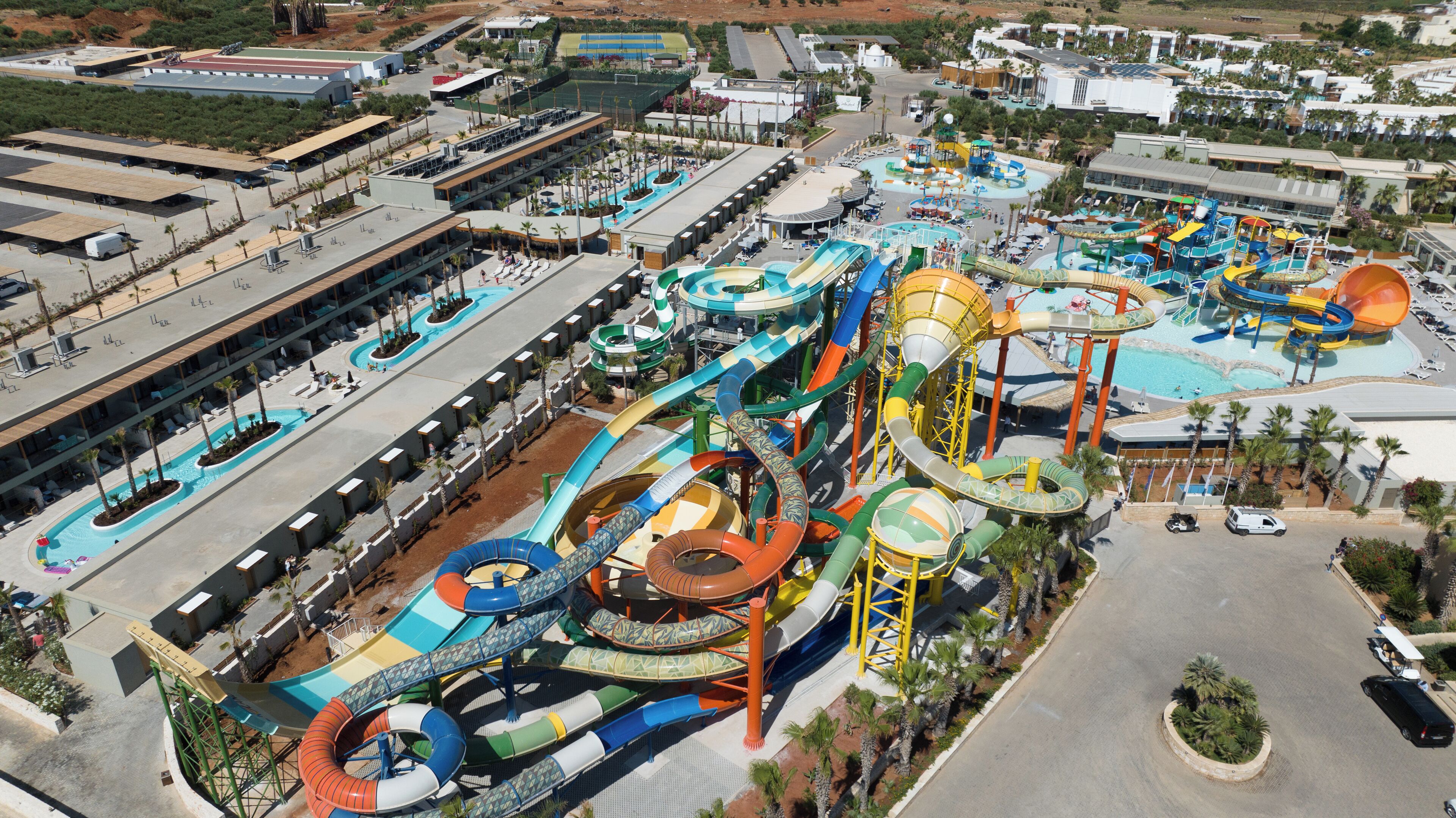 Vista aérea de un parque acuático con varias toboganes y piscinas, rodeado de edificios y palmeras.