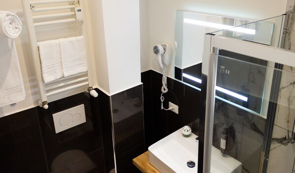 A bathroom with a black tiled wall, a white sink, a glass shower enclosure, and a towel rack with folded towels. - Michelangelo Vatican Rooms (Rome)