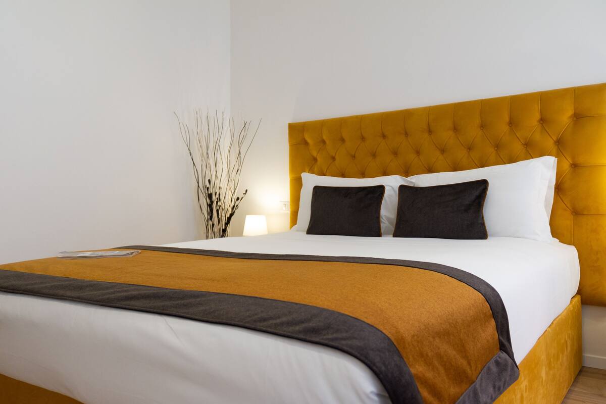 Una cama bien hecha con un cabecero amarillo, almohadas blancas y negras, y una manta marrón.