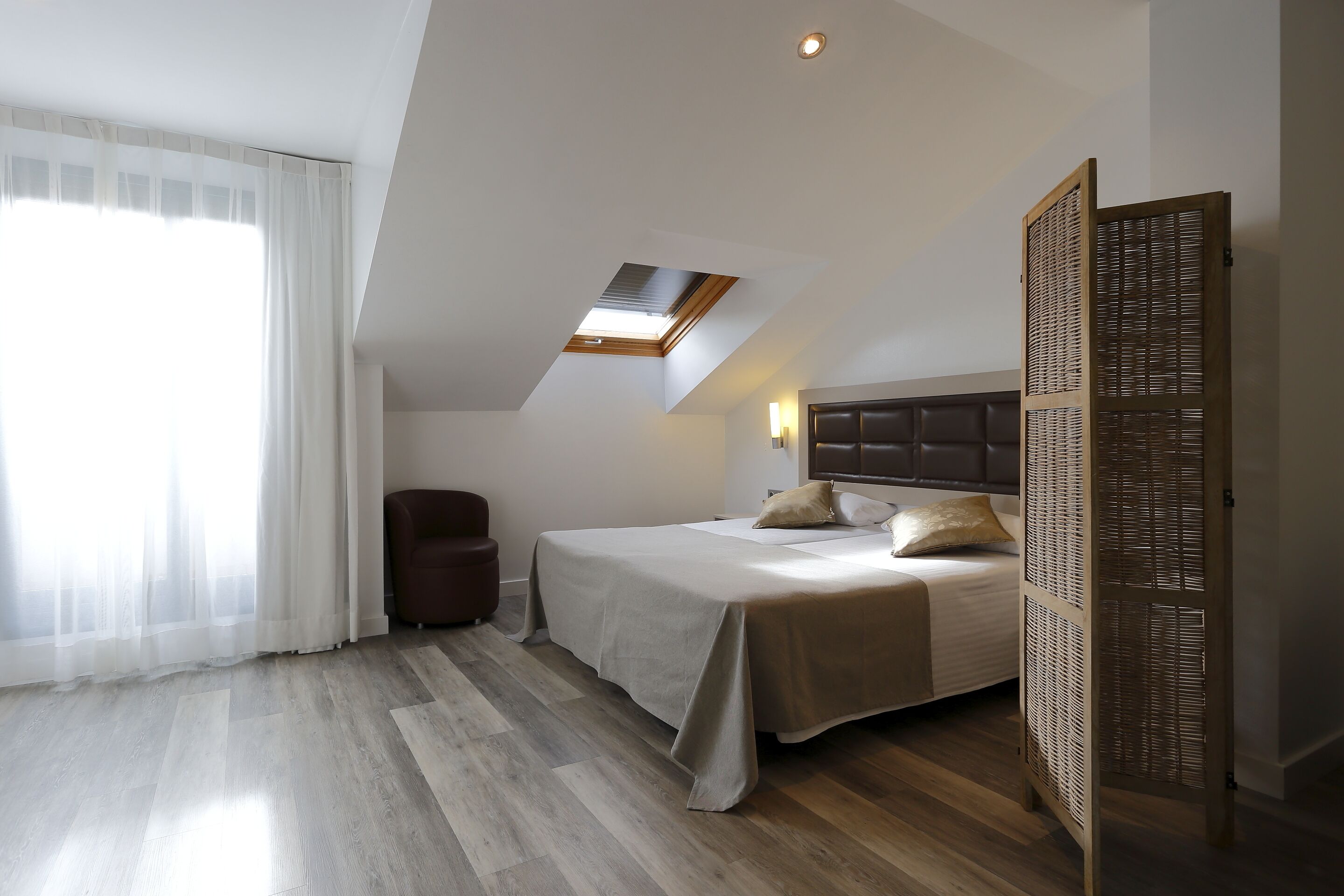 A bedroom with a bed, a chair, a window with sheer curtains, a skylight, and a wooden partition.