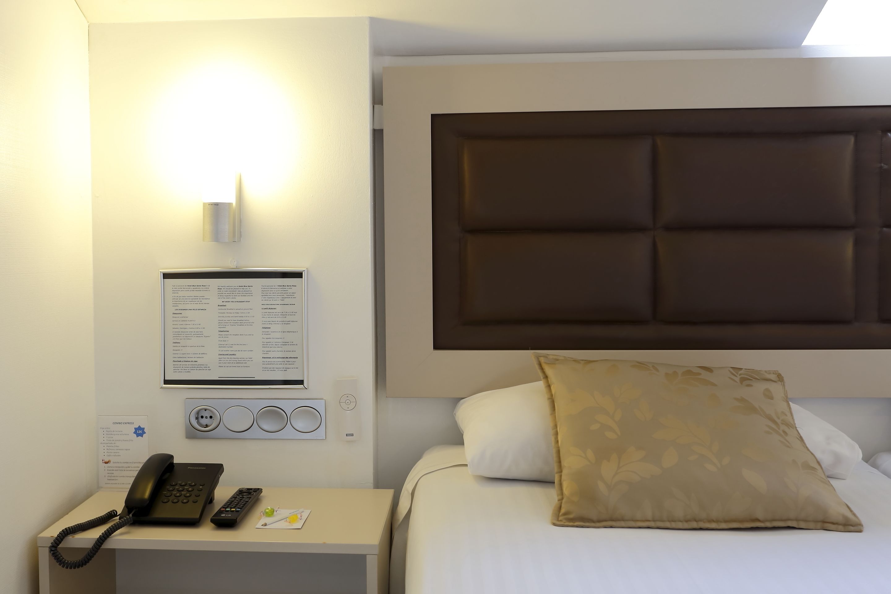 A hotel room with a bed, a bedside table with a phone and remote, a wall-mounted light, and a bulletin board with information.