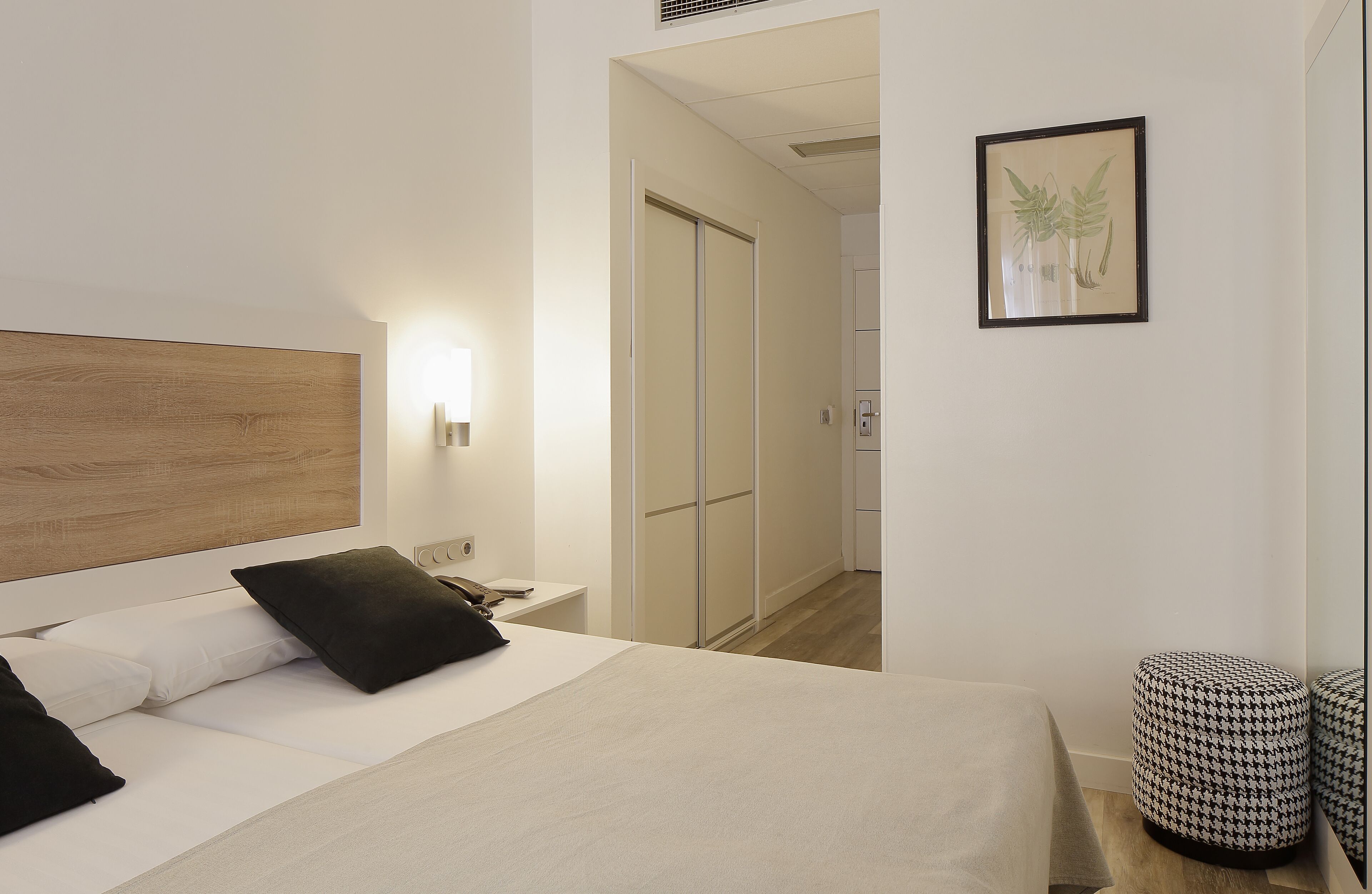 Una habitación de hotel moderna con cabecera de madera, cama con ropa de cama blanca y negra, mesita de noche con lámpara, un grabado botánico enmarcado en la pared y un otomano pequeño y redondo.