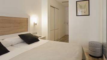 Una habitación de hotel moderna con cabecera de madera, cama con ropa de cama blanca y negra, mesita de noche con lámpara, un grabado botánico enmarcado en la pared y un otomano pequeño y redondo.