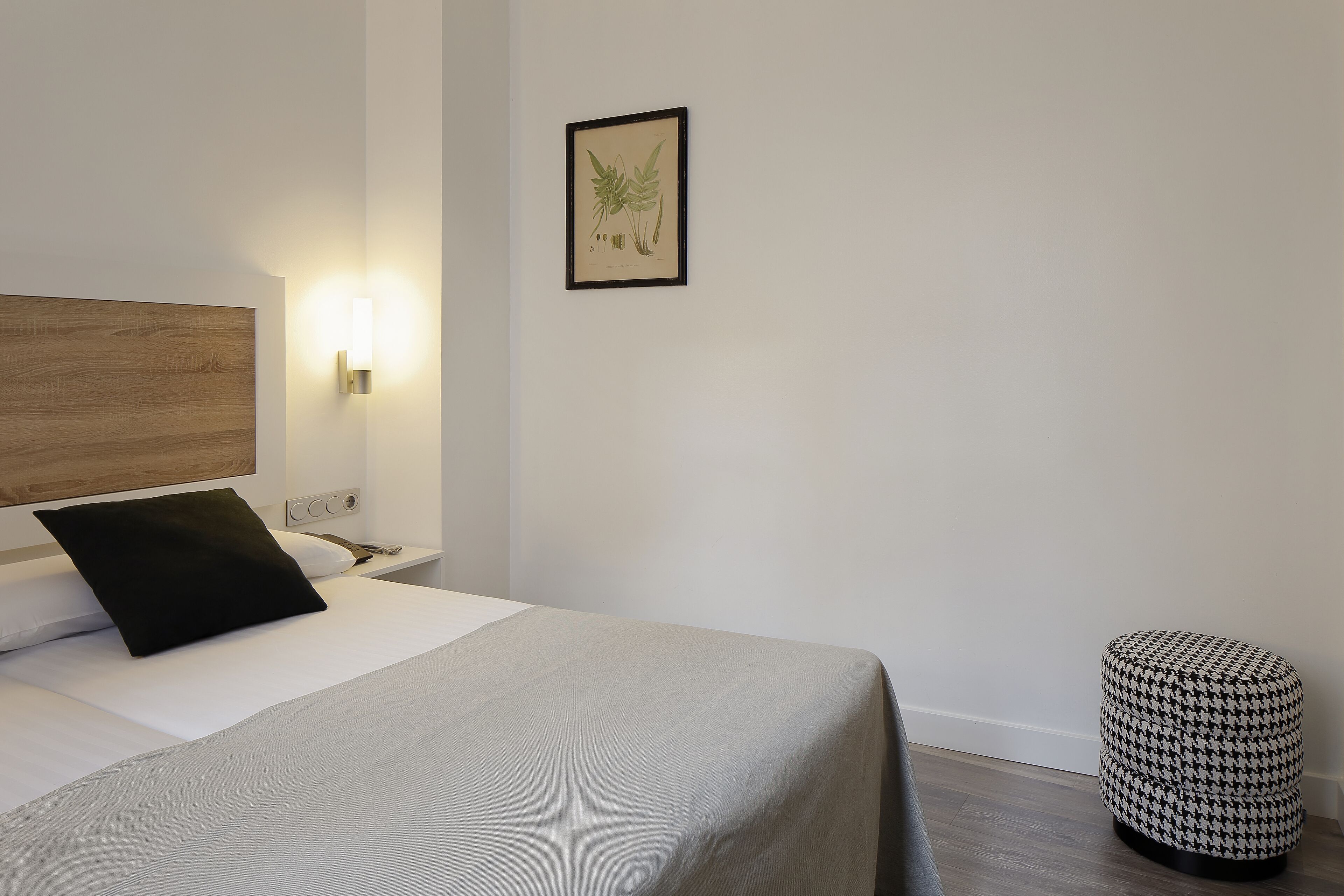 A neatly made bed with a black pillow, a wall-mounted light, and a framed botanical print.