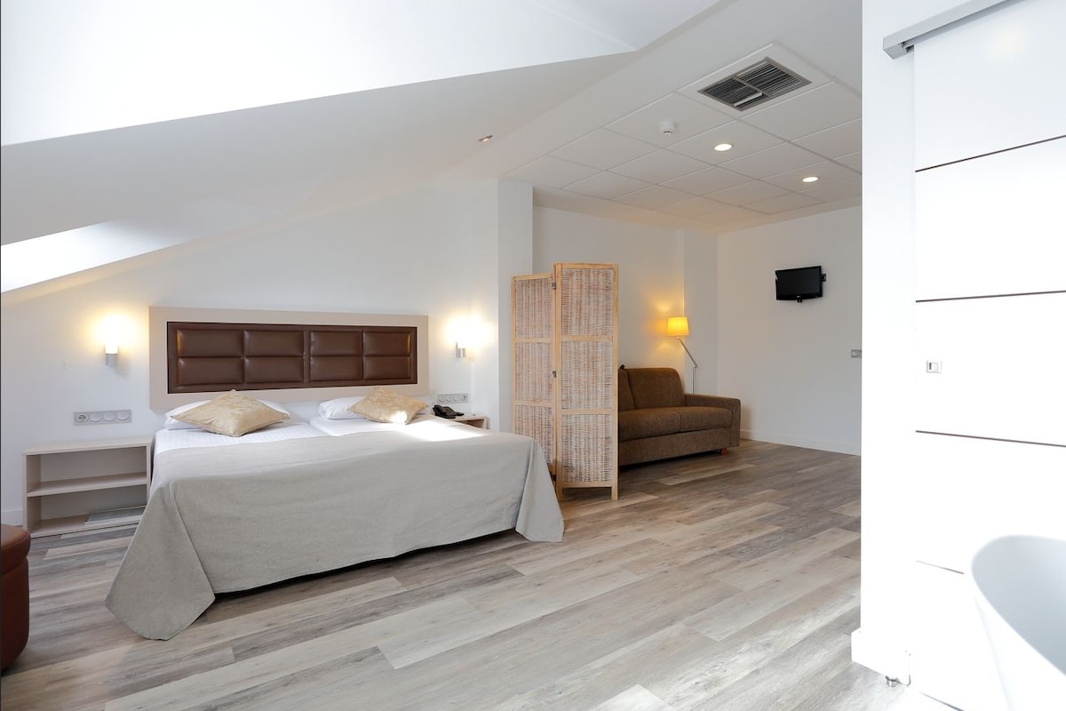 A bedroom with a bed, a chair, a bedside table, a lamp, and a television.