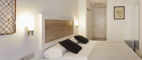 Habitación de hotel con cabecera de madera, dos almohadas negras, cubrecama blanco, mesita de noche con lámpara e impresión botánica enmarcada en la pared.