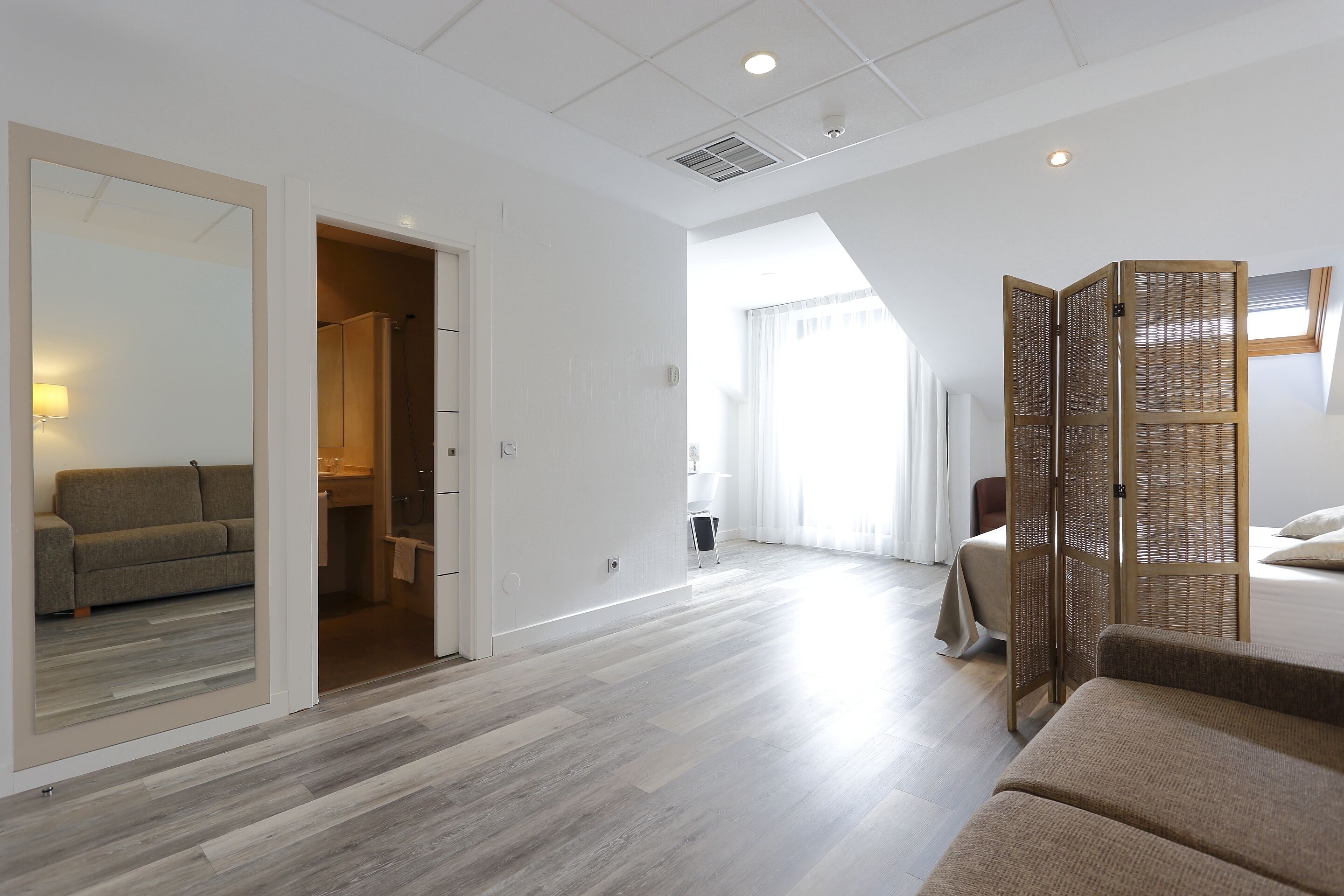 A modern hotel room with a large bed, a sitting area with a sofa, a walk-in closet, and a large window providing natural light.