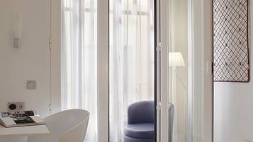 A modern hotel room with a bed, a desk with a chair, a blue armchair, and a sliding glass door leading to a balcony.
