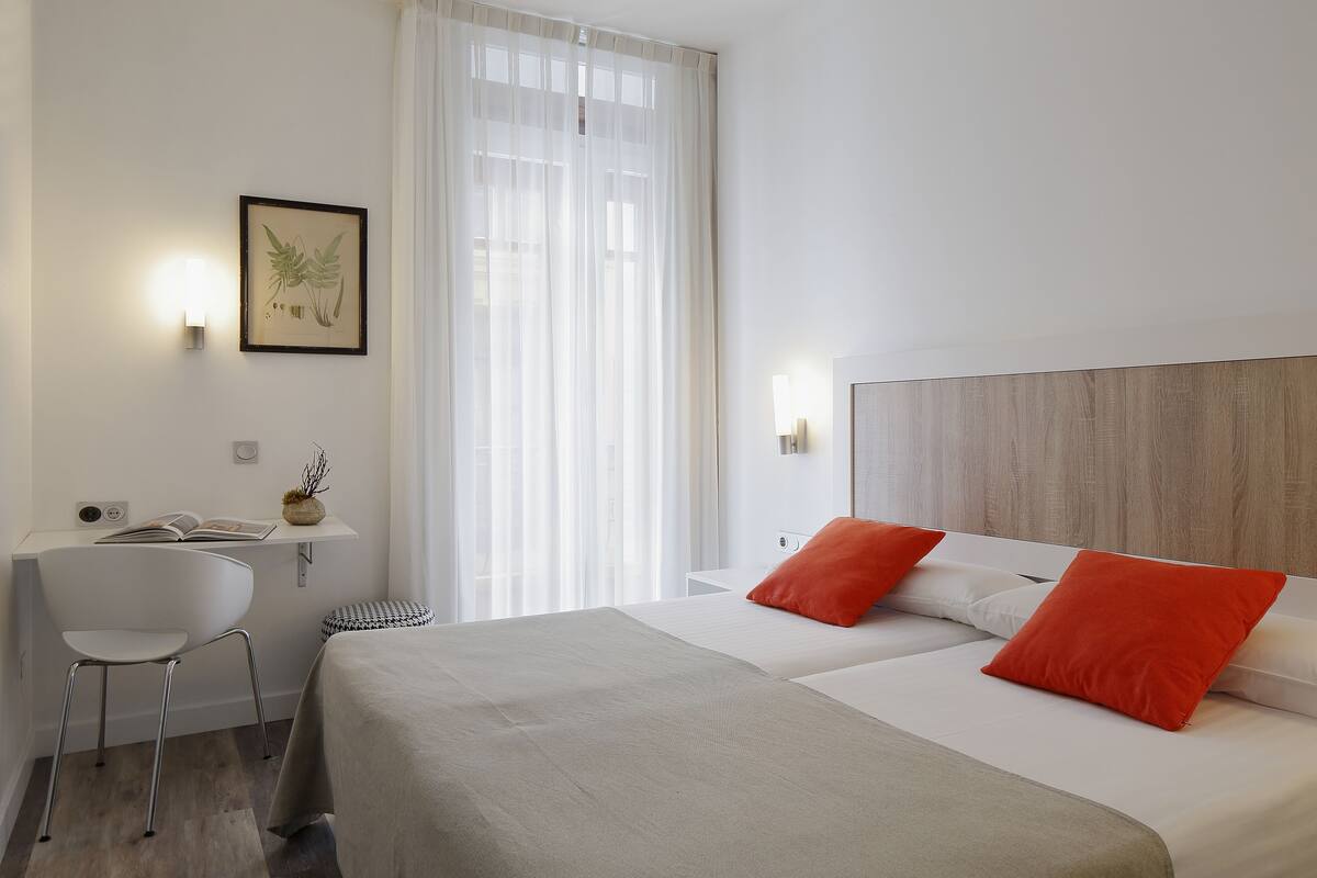 A hotel room with a bed, a desk, a chair, and a window with sheer curtains.