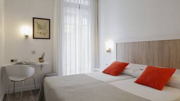 Une chambre d’hôtel avec un lit, un bureau, une chaise et une fenêtre avec des rideaux légers.
