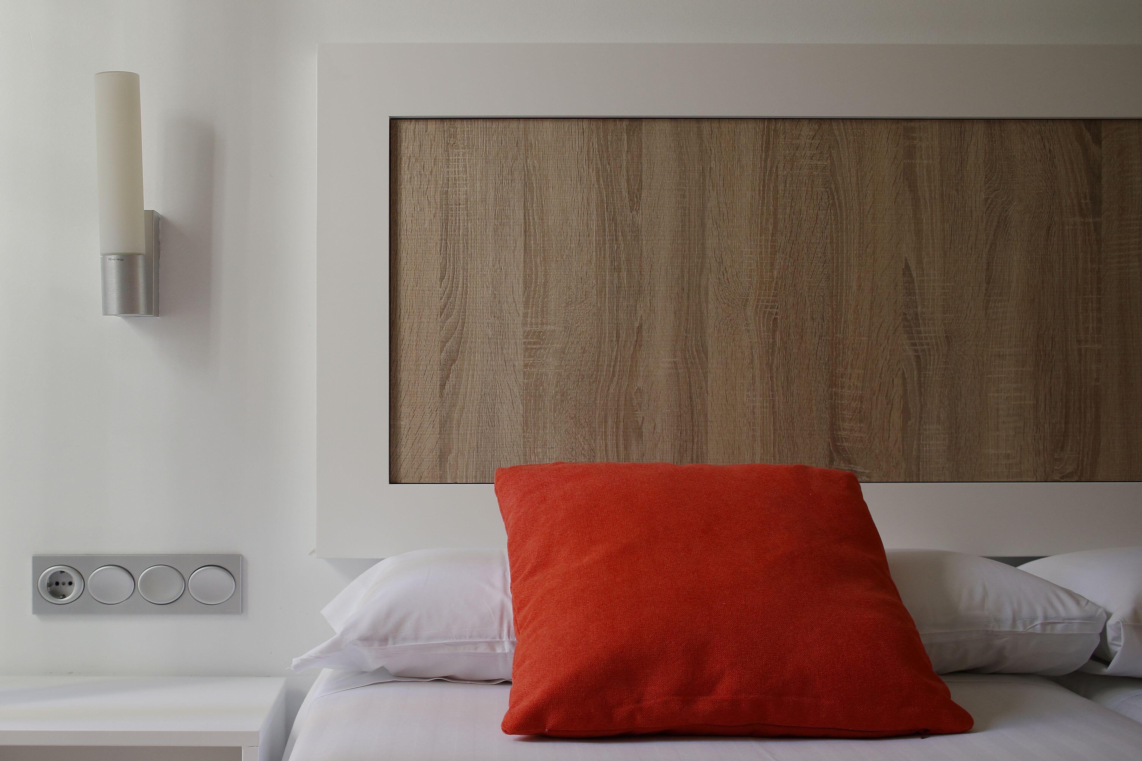 Habitación de hotel con cabecera de madera, mesita de noche con lámpara y almohada roja.