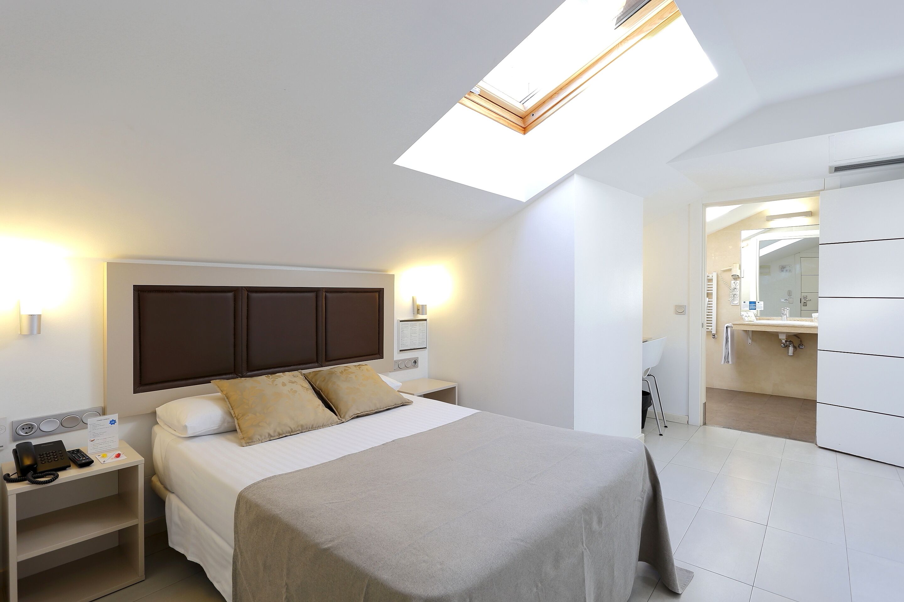 A modern hotel room with a bed, bedside table with a phone, a nightstand, a skylight, and a bathroom with a sink and mirror.