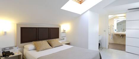 A modern hotel room with a bed, bedside table with a phone, a nightstand, a skylight, and a bathroom with a sink and mirror.