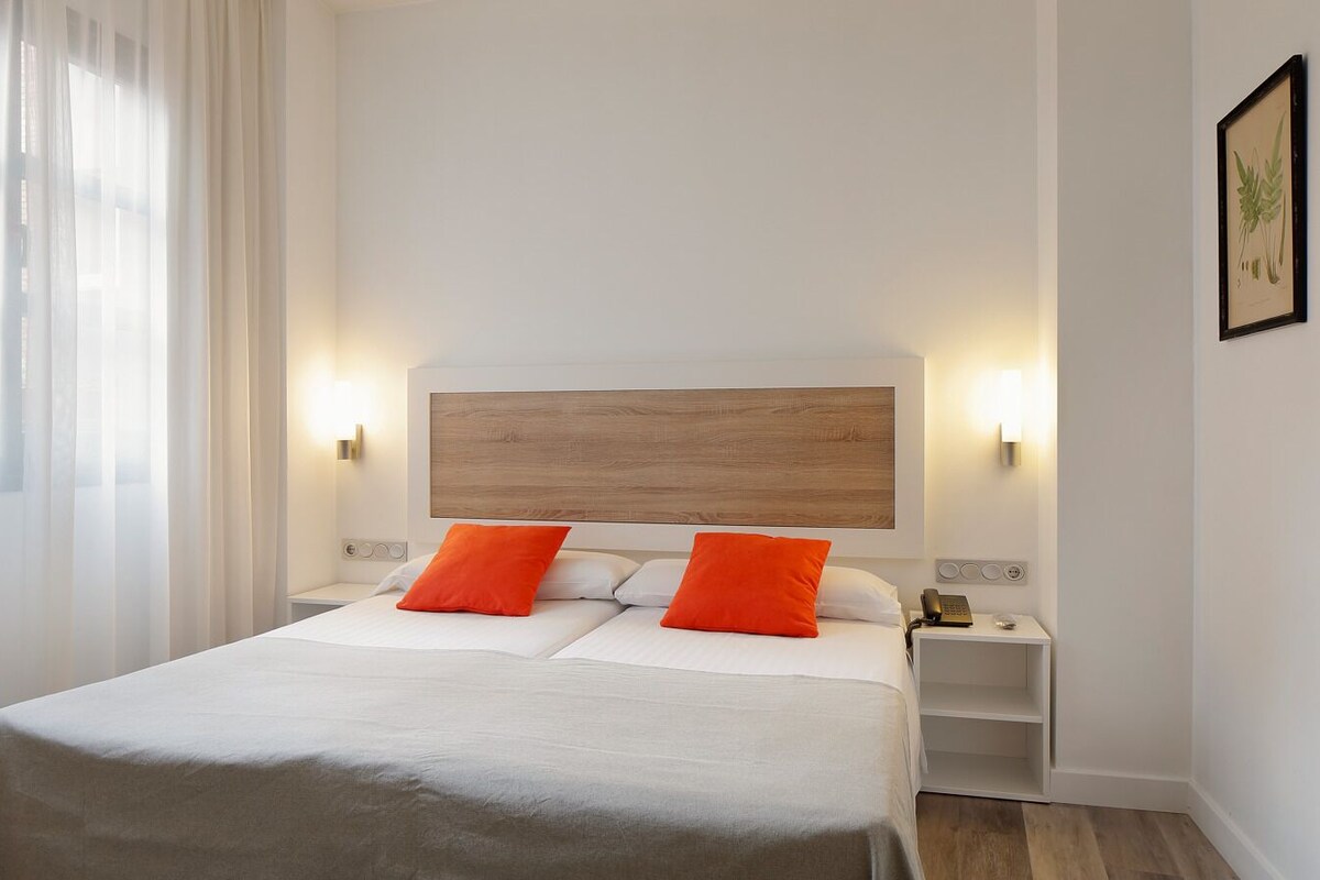 A hotel room with a bed, two orange pillows, a nightstand with a phone, and a wall-mounted light.