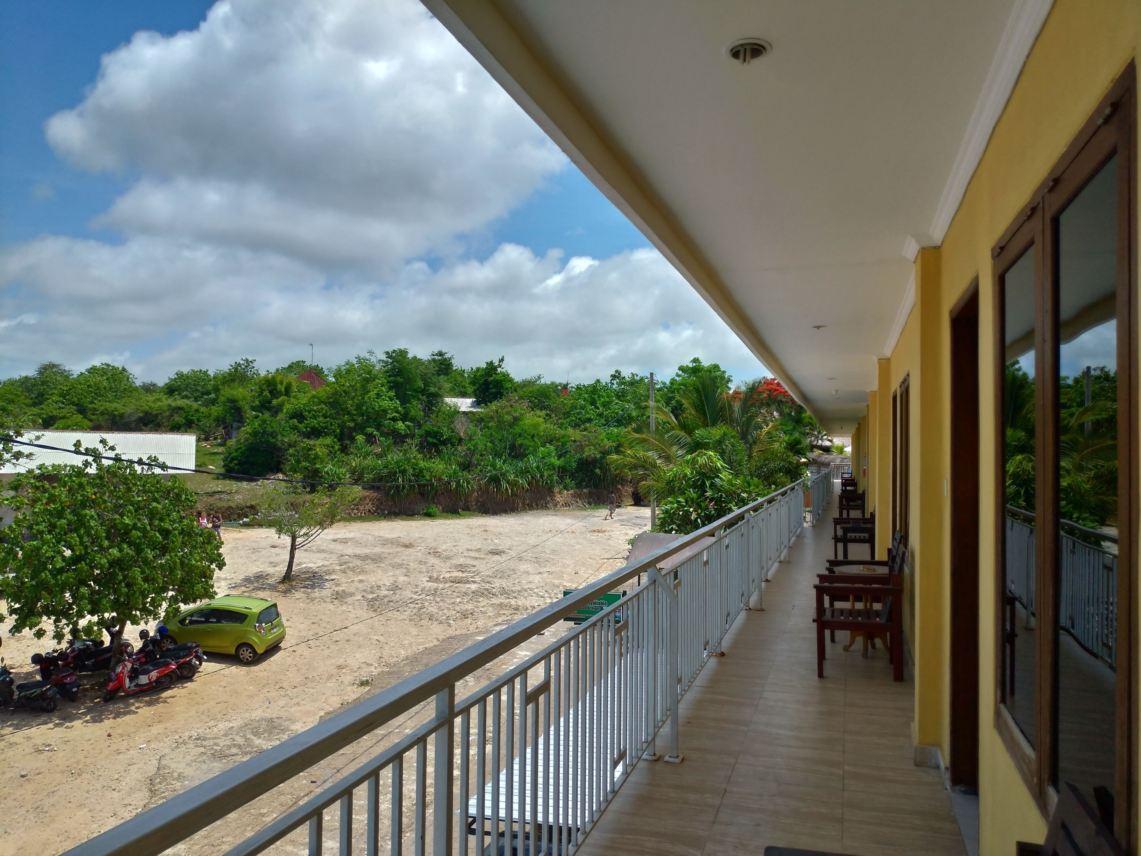 A balcony with a view of a sandy area and greenery, a yellow building, and a clear sky.