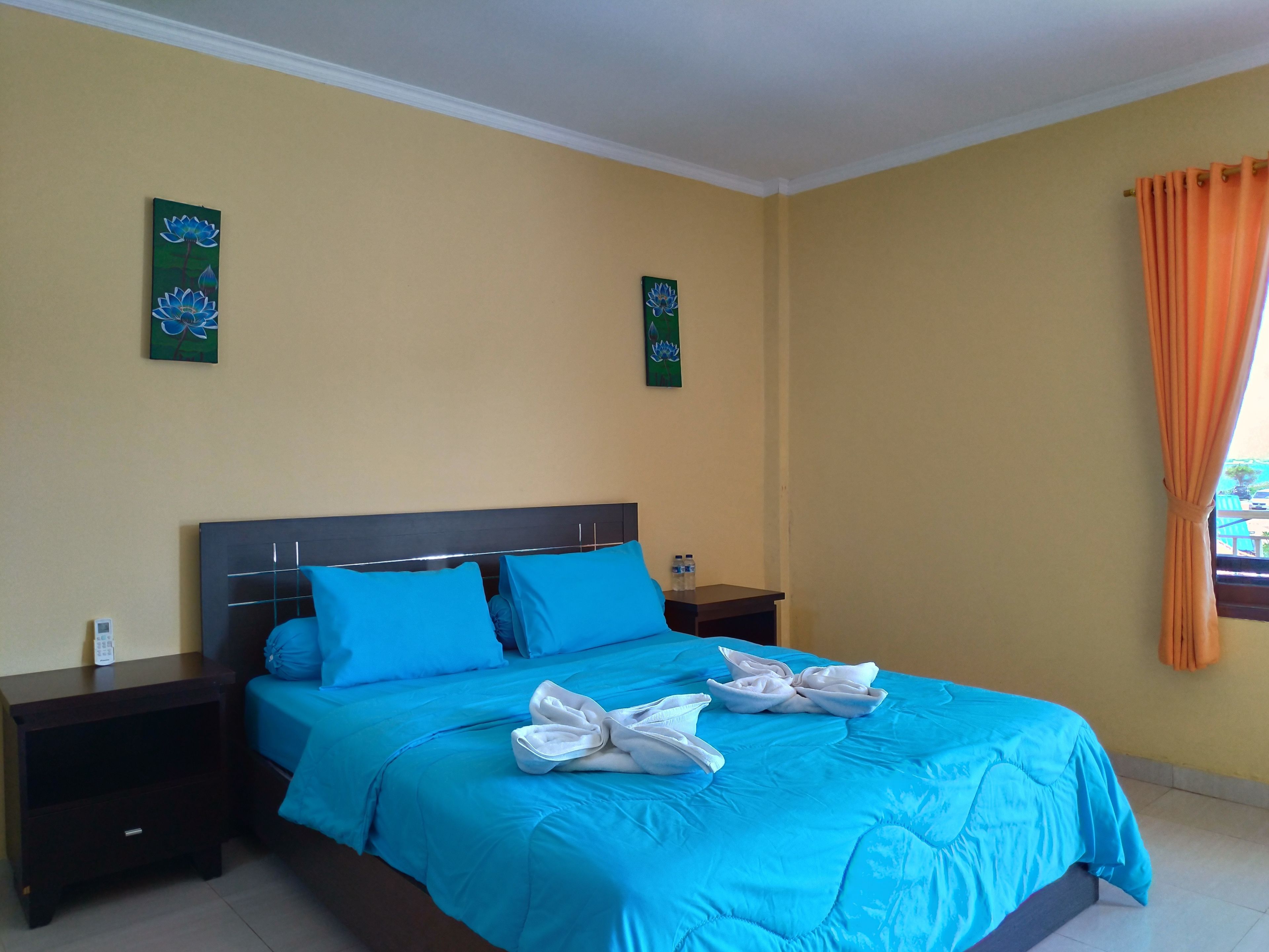 A hotel room with a bed, two nightstands, a window with orange curtains, and two wall art pieces.