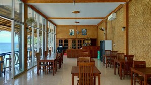 A spacious dining area with wooden tables and chairs, large windows offering a view of the ocean, and a bar area with a counter and seating. - Balangan Paradise Hostel and Restaurant (Jimbaran)
