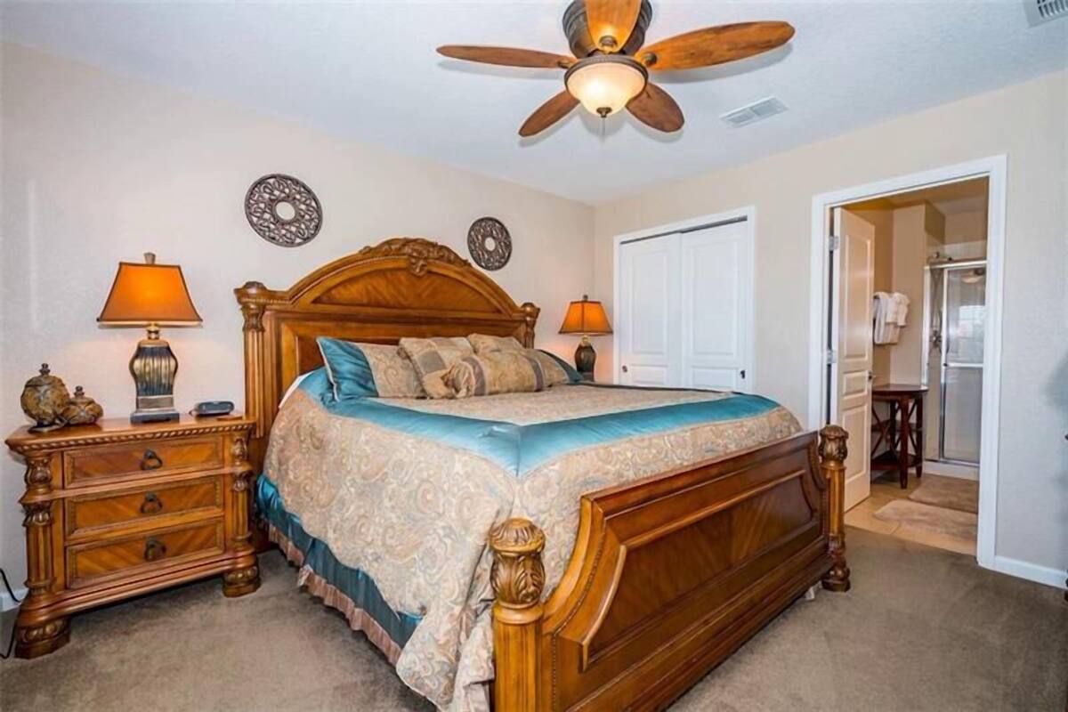 A bedroom with a wooden bed, two bedside tables with lamps, a ceiling fan, and a door leading to a bathroom.