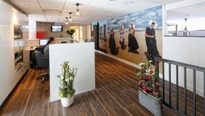 Un área de recepción moderna con piso de madera, una mesa de recepción, un gran mural con escena de playa y plantas en macetas.